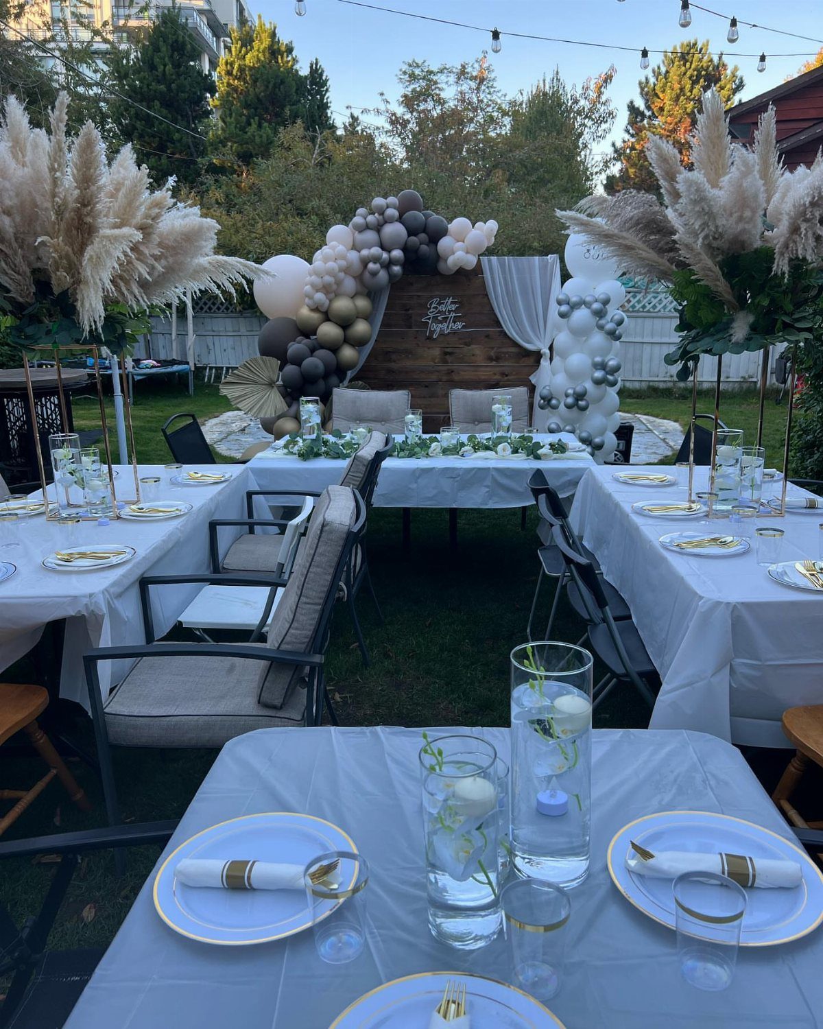 Outdoor party setup with white tables, gold plates, and balloon arch in a backyard.