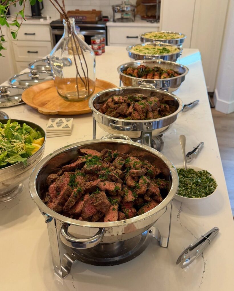 Sliced grilled meats in silver chafing dishes on a white buffet table.