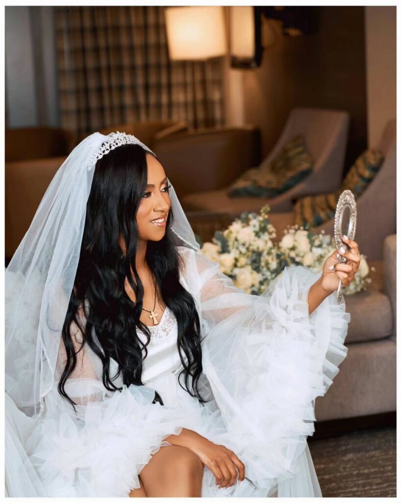 Bride in a white robe and veil looking into a small hand mirror.