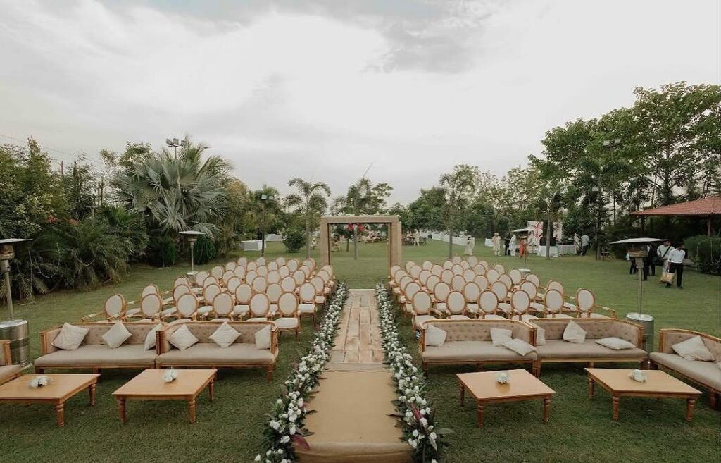 Outdoor wedding ceremony setup with beige couches, wooden tables, and rows of chairs.