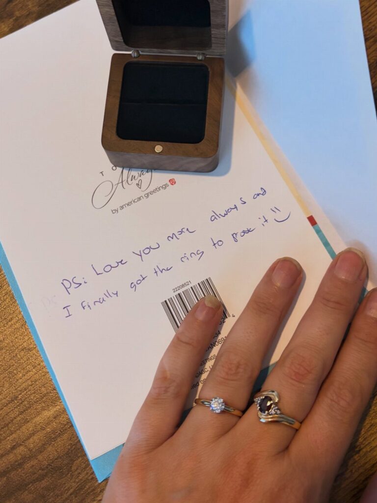 Hand wearing engagement rings resting on a handwritten love note and ring box.