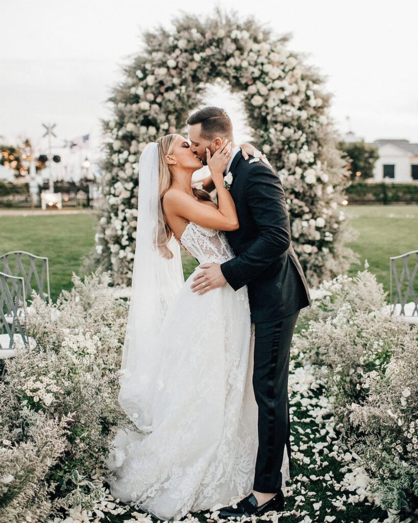 Bride and groom kissing in front of a large white floral wedding arch.