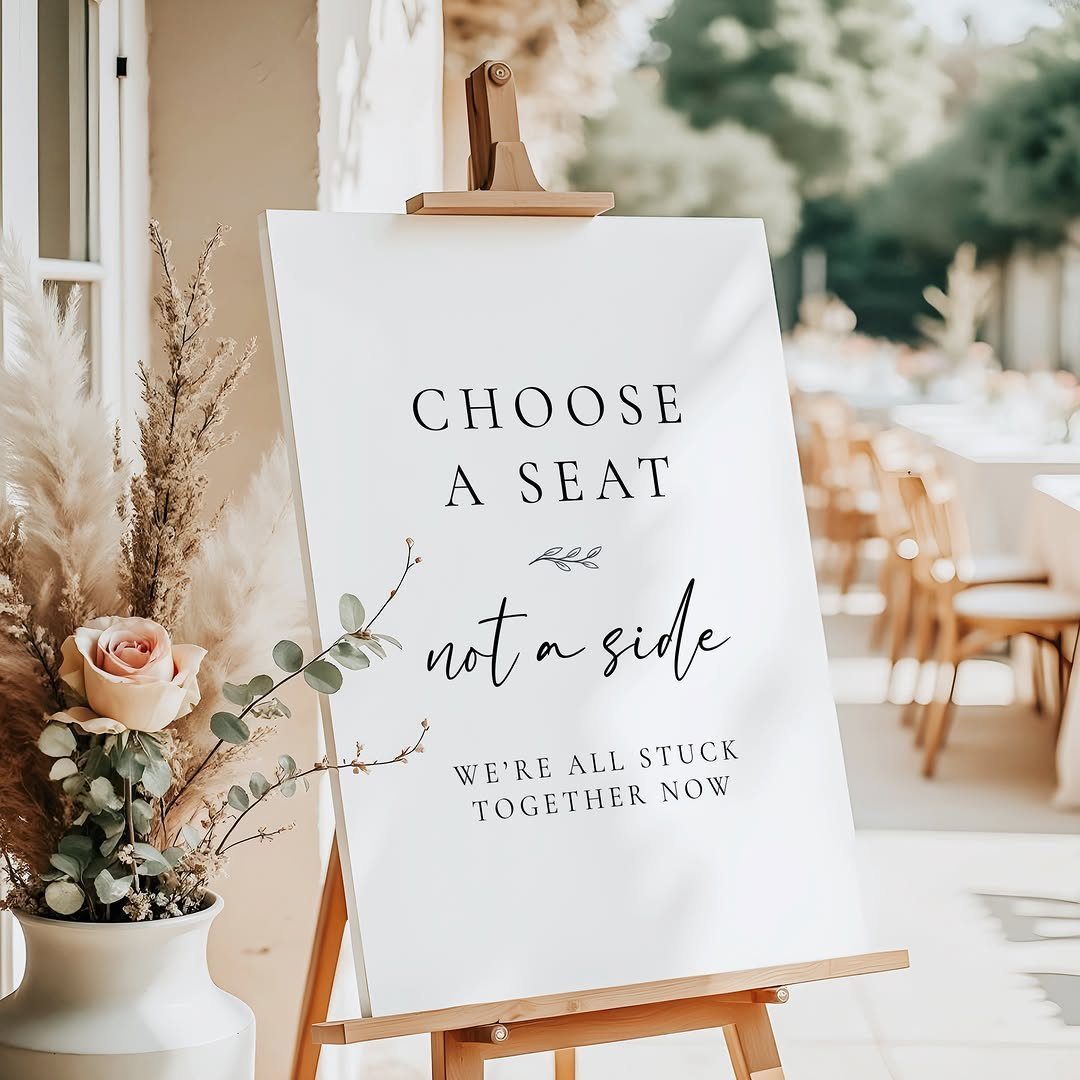 White wedding sign on a wooden easel with humorous seating instructions and floral accents.