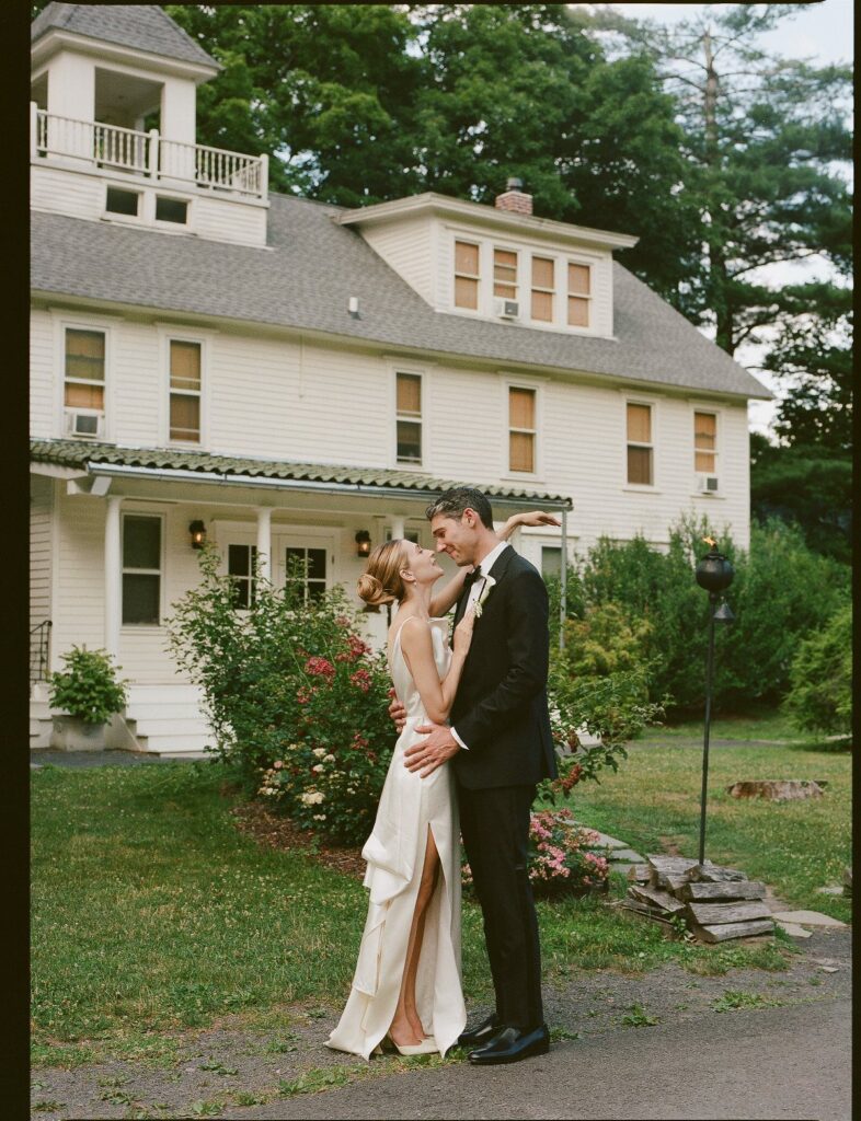 Bride and groom embracing in front of a white farmhouse in a rural setting.
