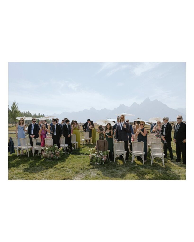 Wedding guests seated in white chairs on a grassy field with mountains behind.