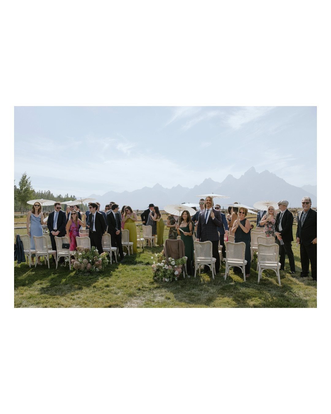 Wedding guests seated in white chairs on a grassy field with mountains behind.