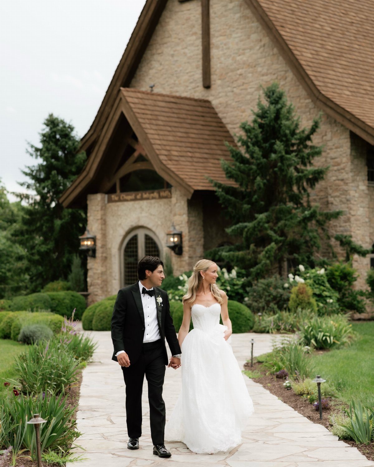 Bride and groom holding hands walking in front of a stone wedding chapel.