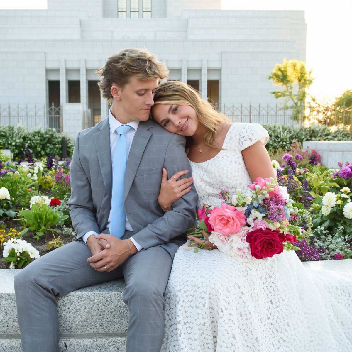 Bride in lace dress and groom in grey suit posing before a temple.