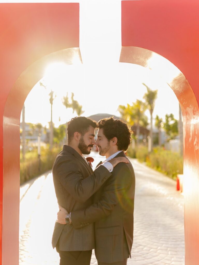 Two men in grey suits embracing under a red arch in sunny Los Cabos.