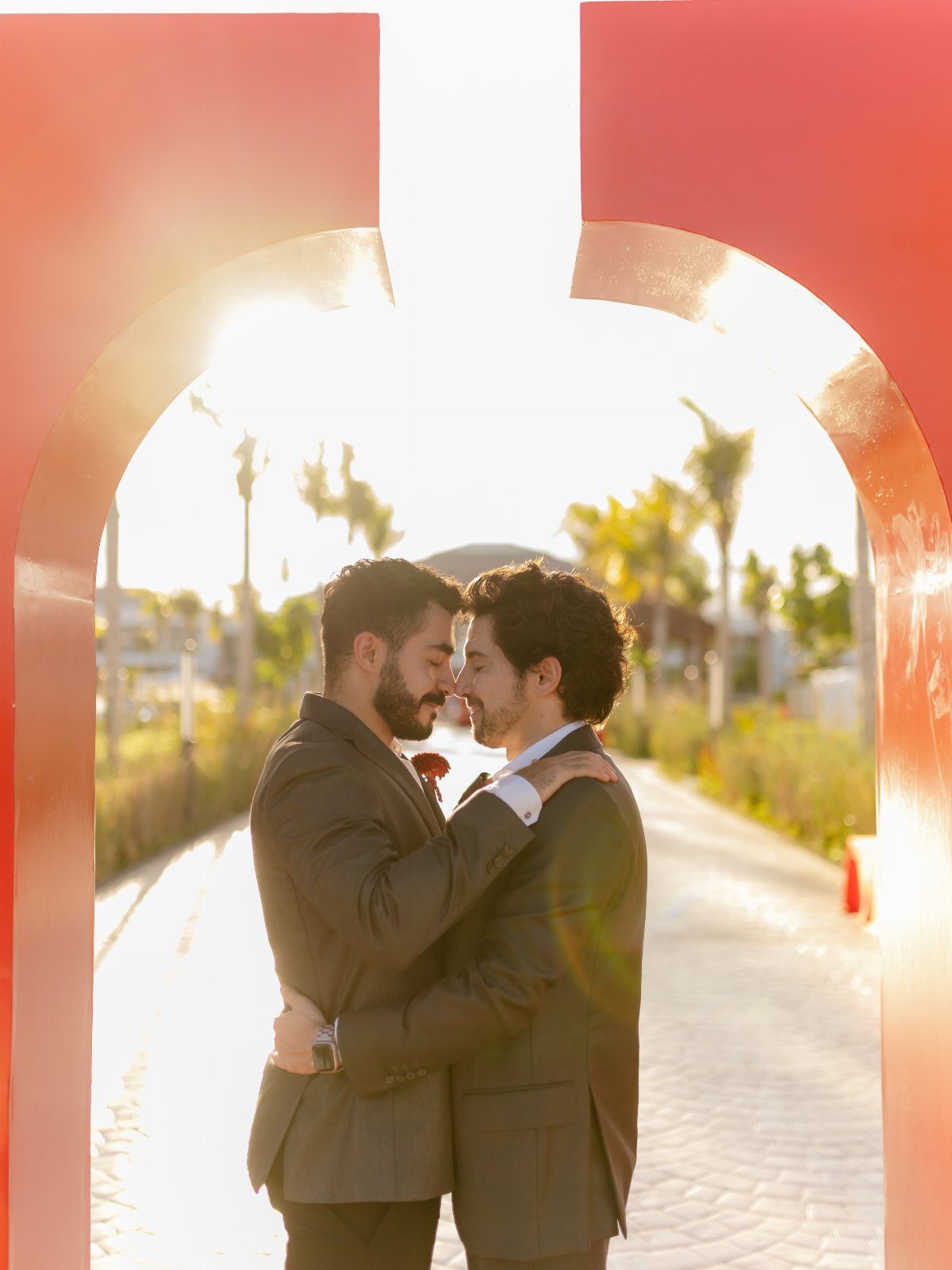Two men in grey suits embracing under a red arch in sunny Los Cabos.