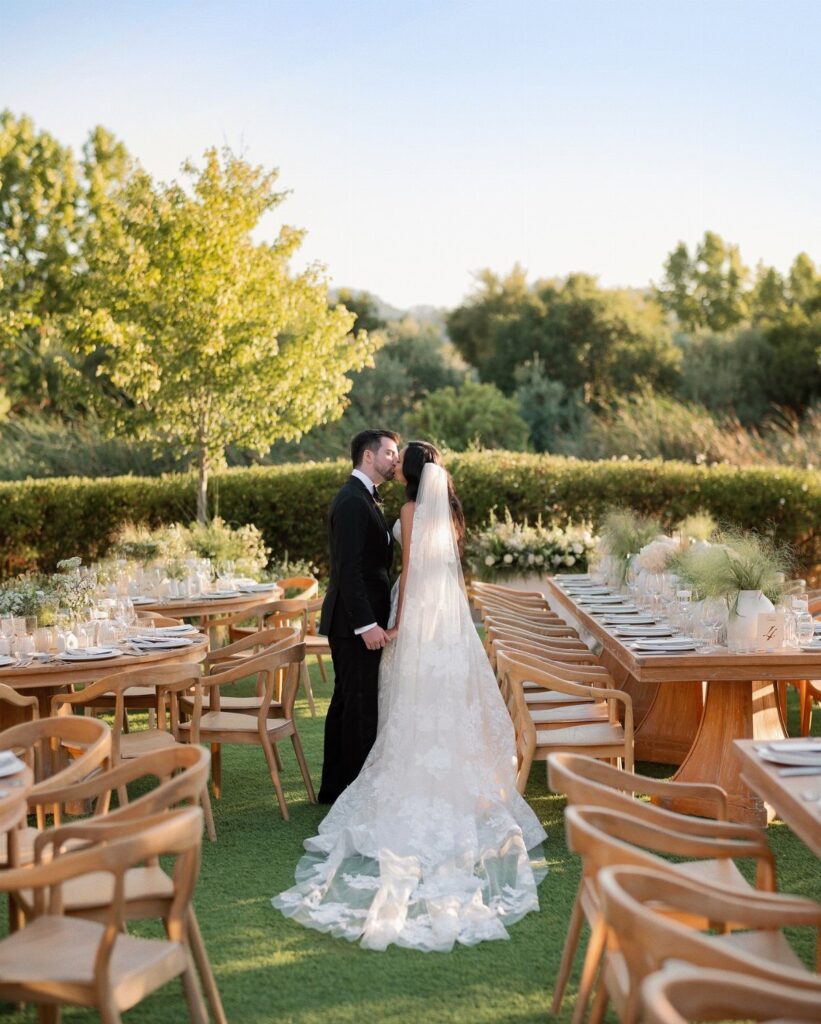 Bride and groom kissing at an outdoor wedding reception with long wooden tables.