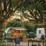 Green food truck and white trailer under glowing string lights in a garden