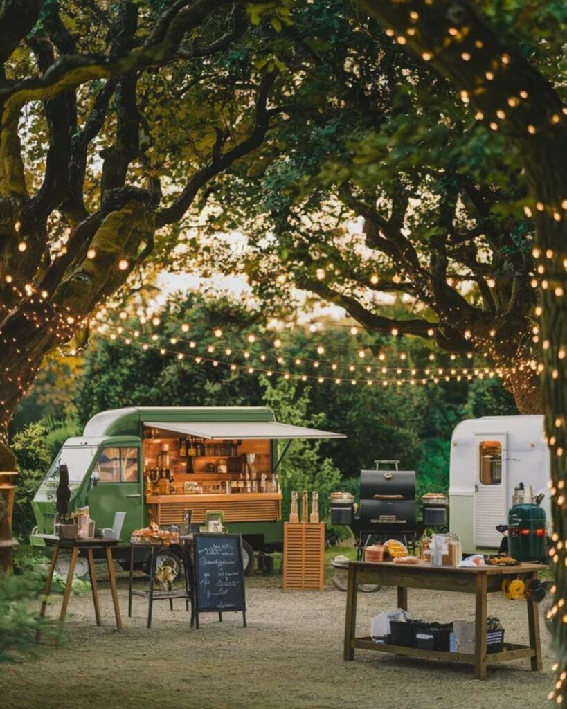 Green food truck and white trailer under glowing string lights in a garden