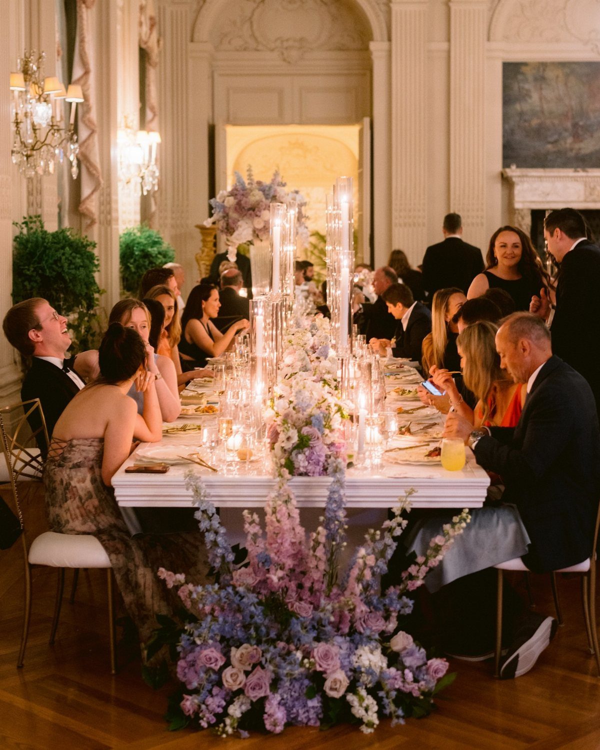 Wedding guests seated at a long table with purple floral arrangements and candles.