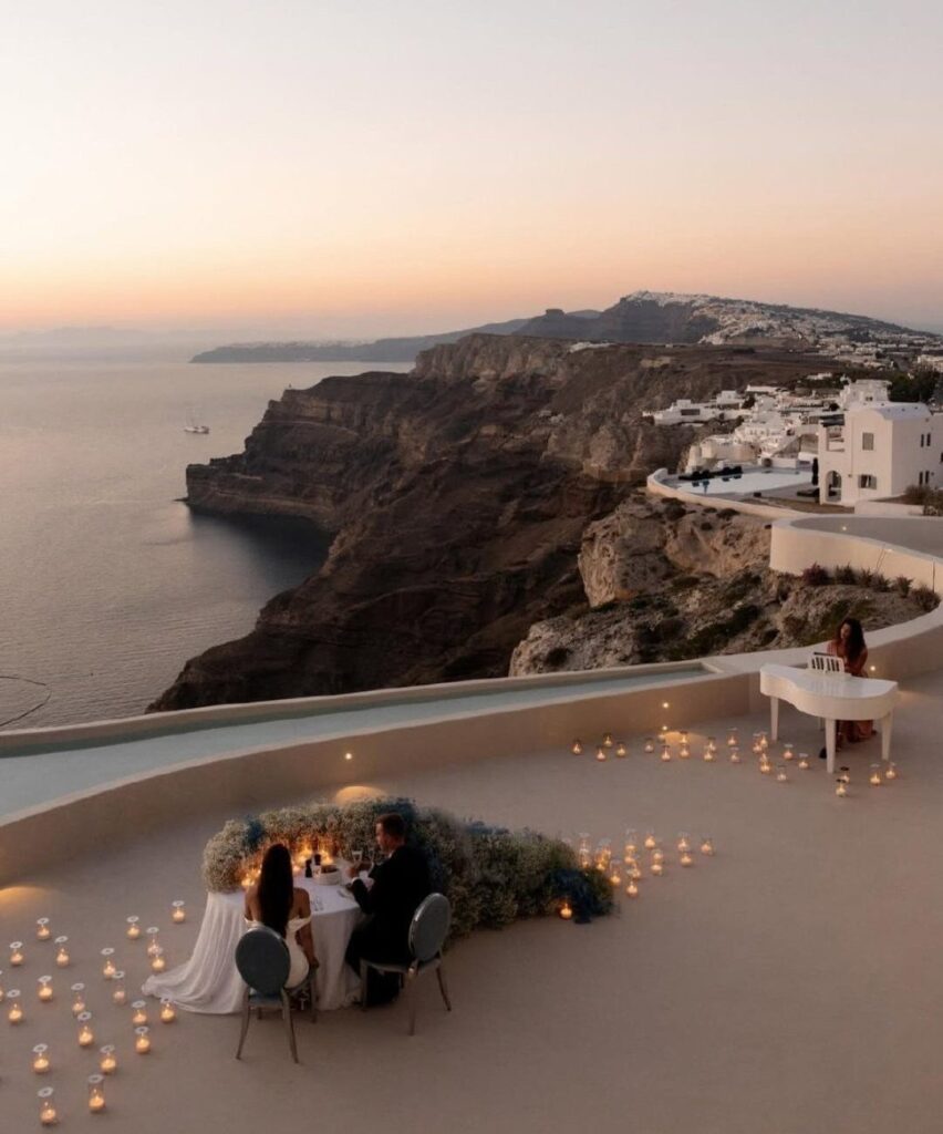 Wedding couple dining at a candlelit table on a Santorini cliffside terrace at sunset.