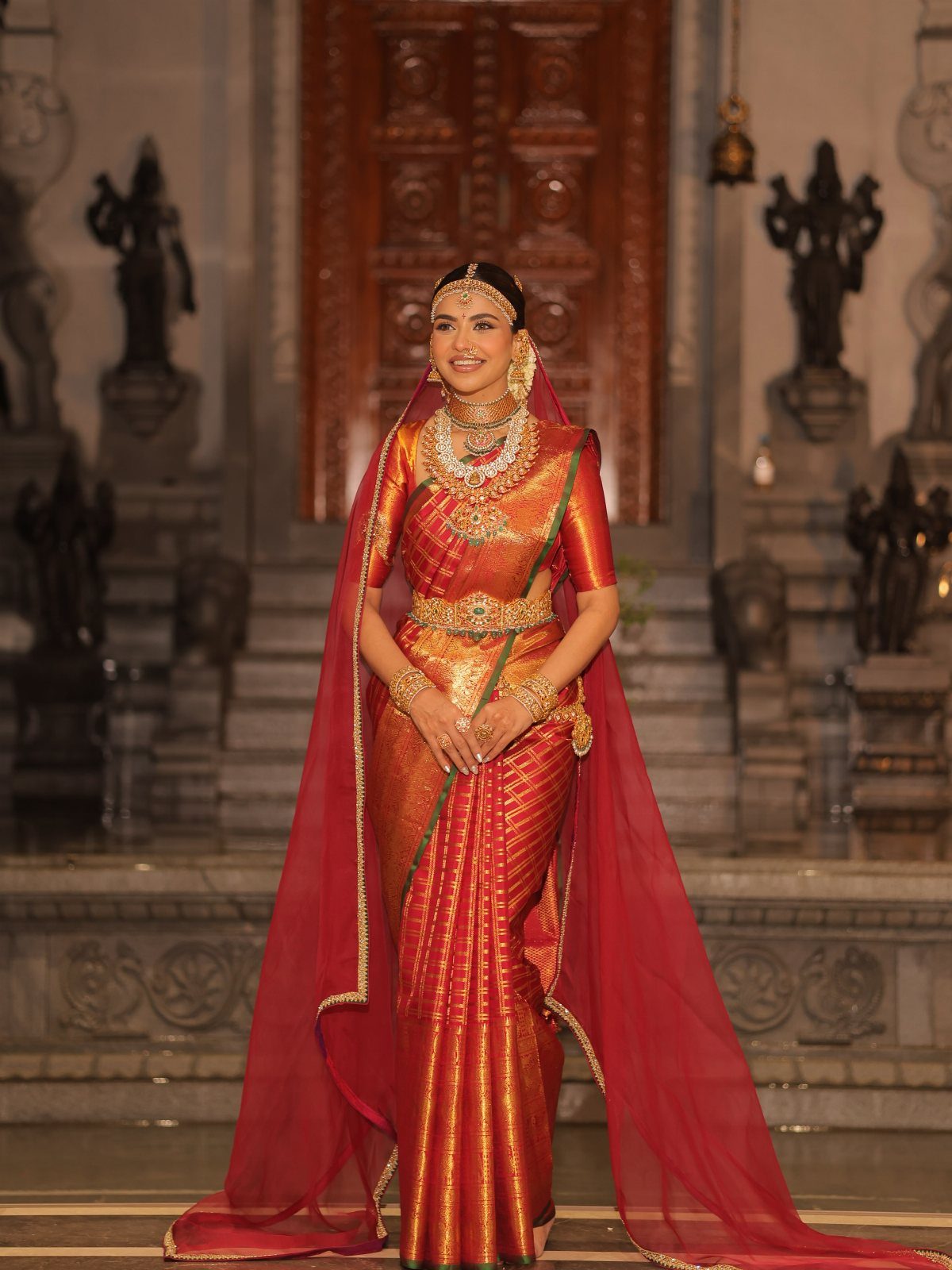 South Indian bride in red silk saree and gold jewelry standing in temple