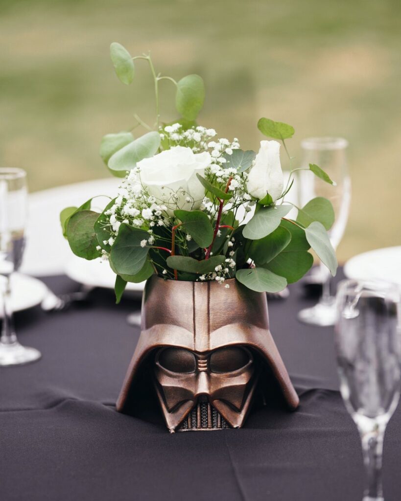 Darth Vader helmet vase holding white roses and greenery on a black table