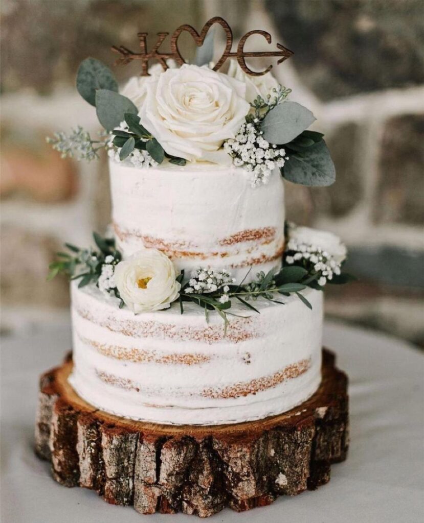 Two-tier naked wedding cake with white flowers on a rustic wooden tree slice base.