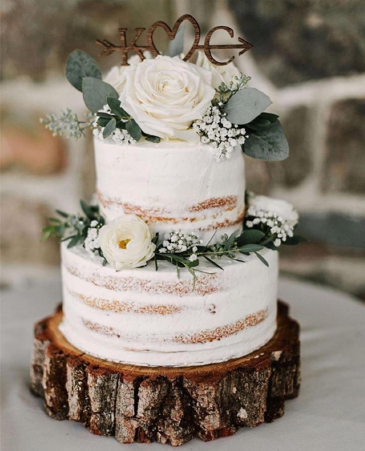Two-tier naked wedding cake with white flowers on a rustic wooden tree slice base.