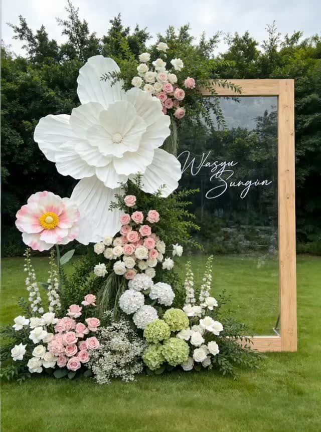 Wooden mirror frame decorated with giant paper flowers and fresh floral arrangements outdoors.