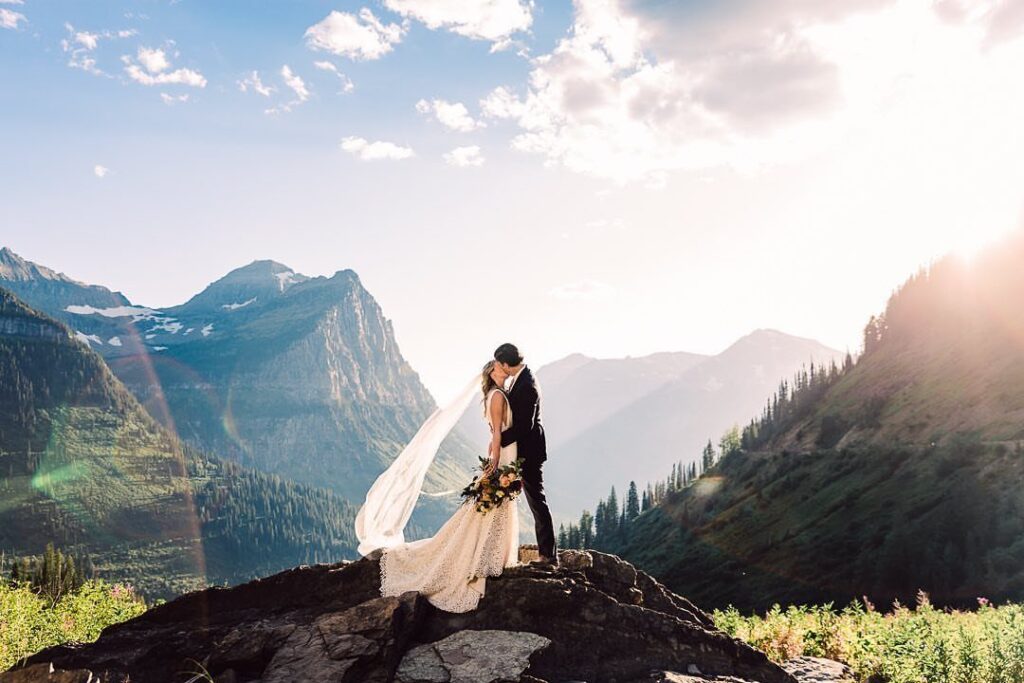 Bride and groom kissing on a rocky peak overlooking a mountainous forest valley.