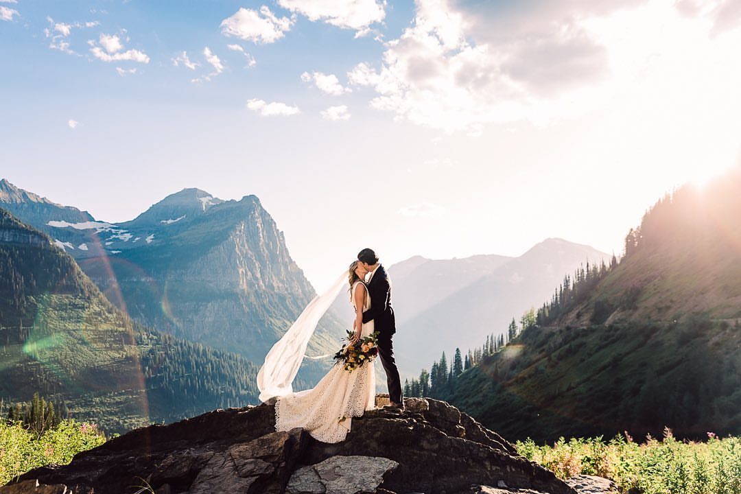 Bride and groom kissing on a rocky peak overlooking a mountainous forest valley.