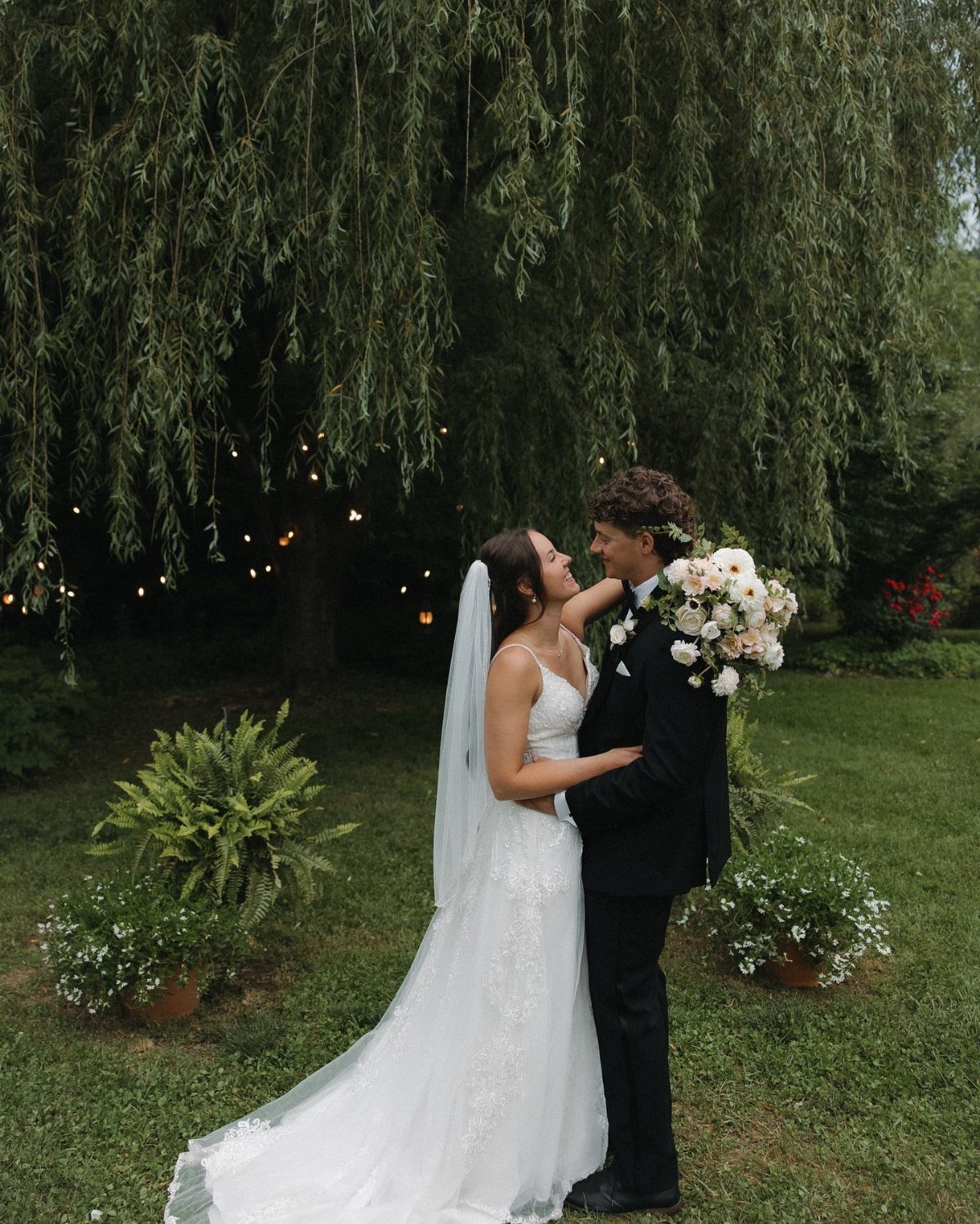 Bride and groom embracing under the drooping branches of a large willow tree.